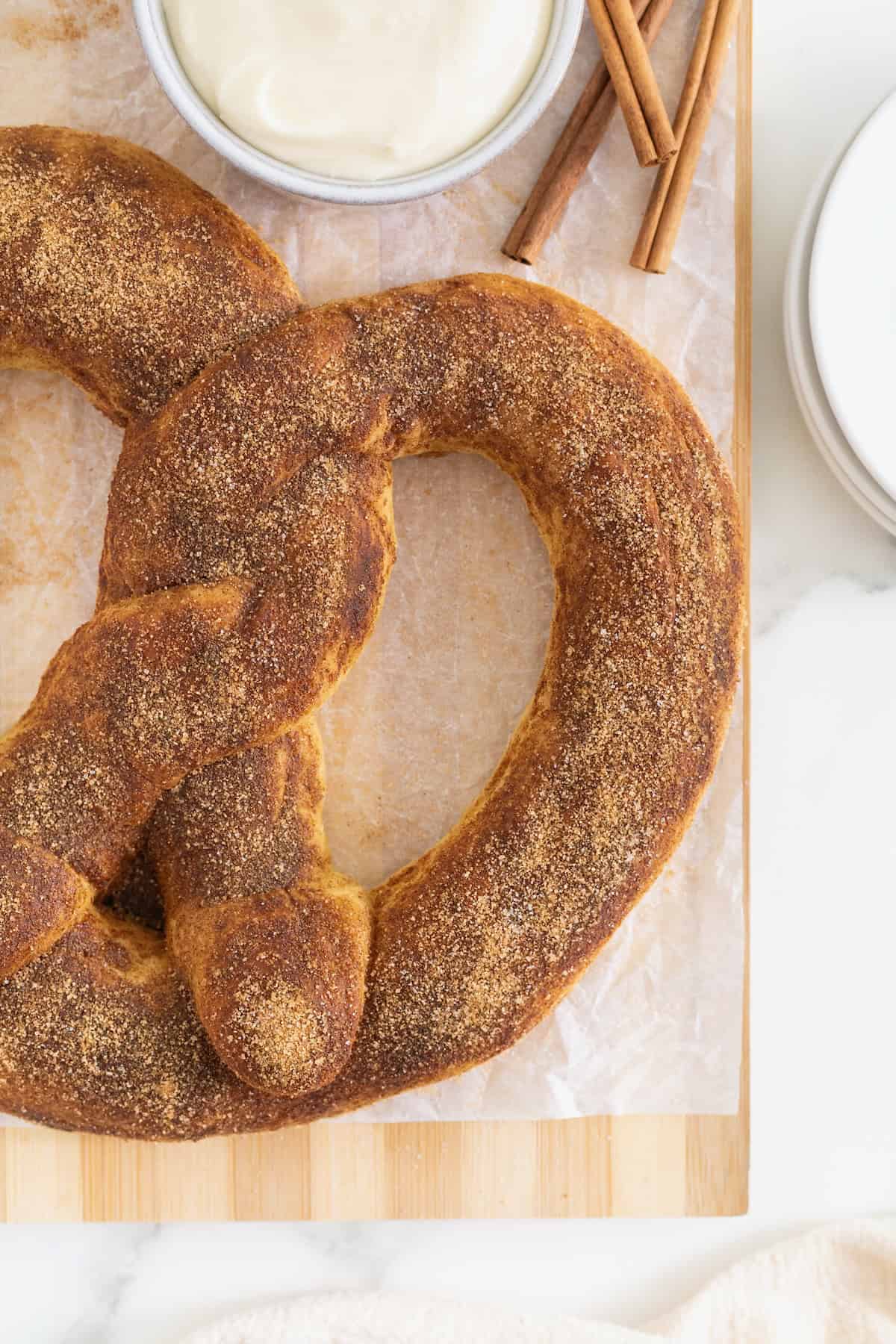 A jumbo sized soft pretzel covered in cinnamon sugar on a parchment lined wood cutting board. Next to the board are a few cinnamon sticks.