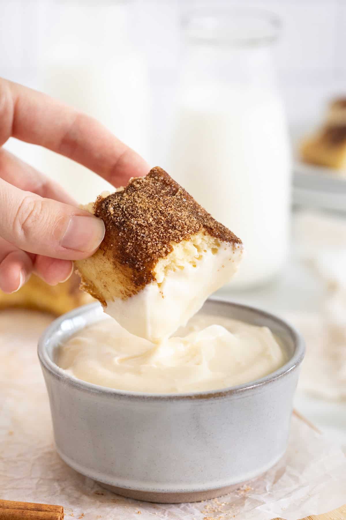 A piece of soft cinnamon sugar pretzel being dipped into a small dish of cream cheese frosting.