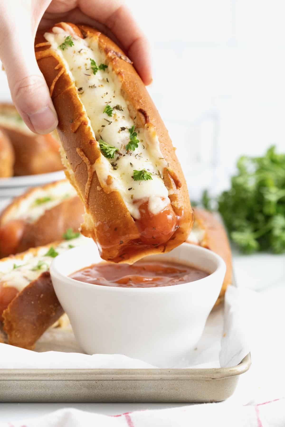 A hand dipping a cheesy garlic bread hot dog into a small white container of marinara sauce.
