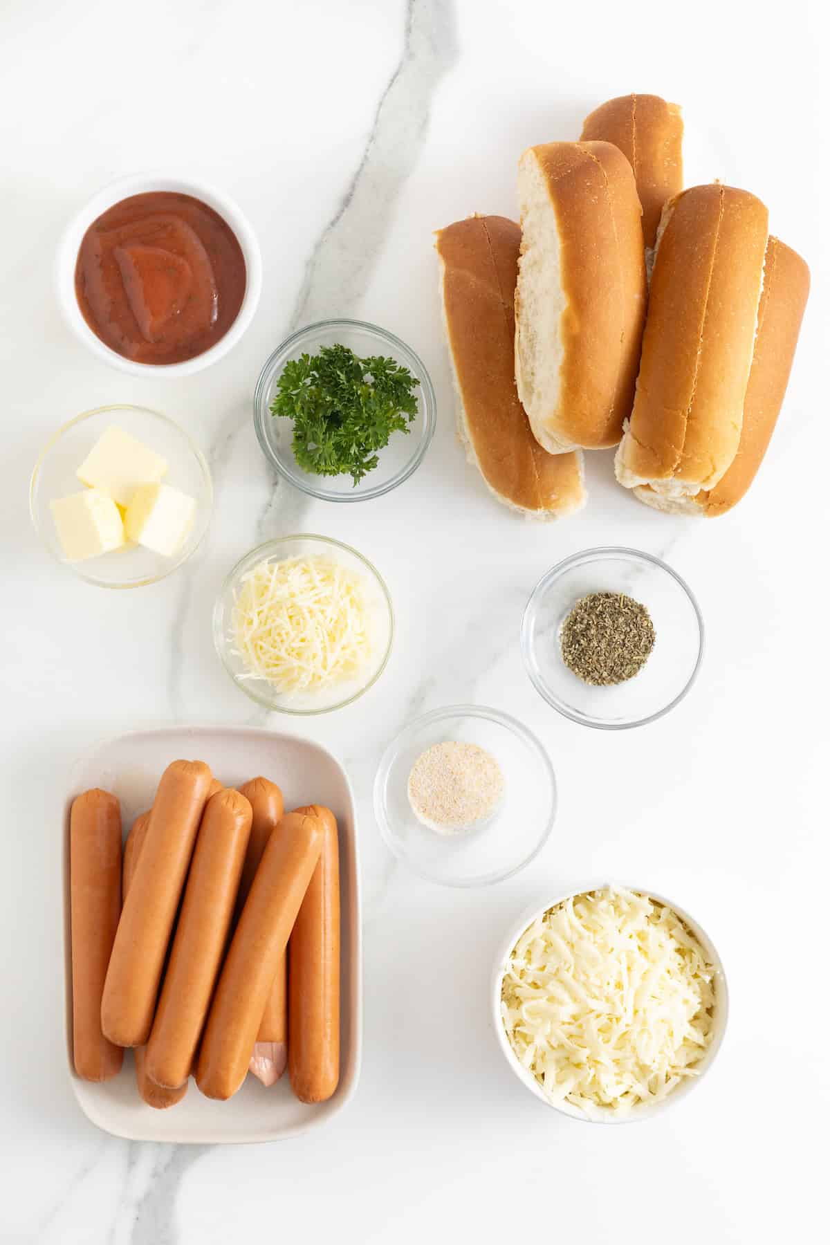 Ingredients to make cheesy garlic bread hot dogs in small glass containers on a white marble counter.