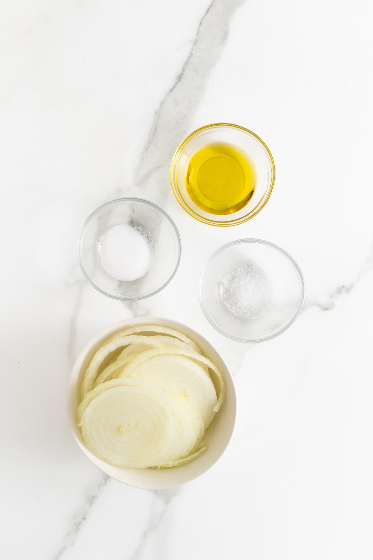 Ingredients needed to caramelize onions in small glass dishes on a white marble counter.