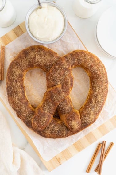A jumbo sized soft pretzel covered in cinnamon sugar on a parchment lined wood cutting board. Next to the board are a few cinnamon sticks.