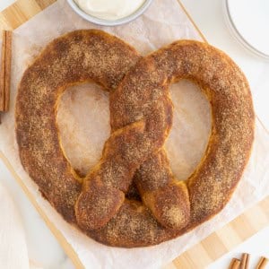 A jumbo sized soft pretzel covered in cinnamon sugar on a parchment lined wood cutting board. Next to the board are a few cinnamon sticks.