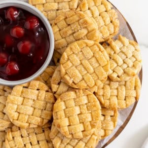 Lattice pie cookies on a serving plate with a small white bowl of cherry pie filling.