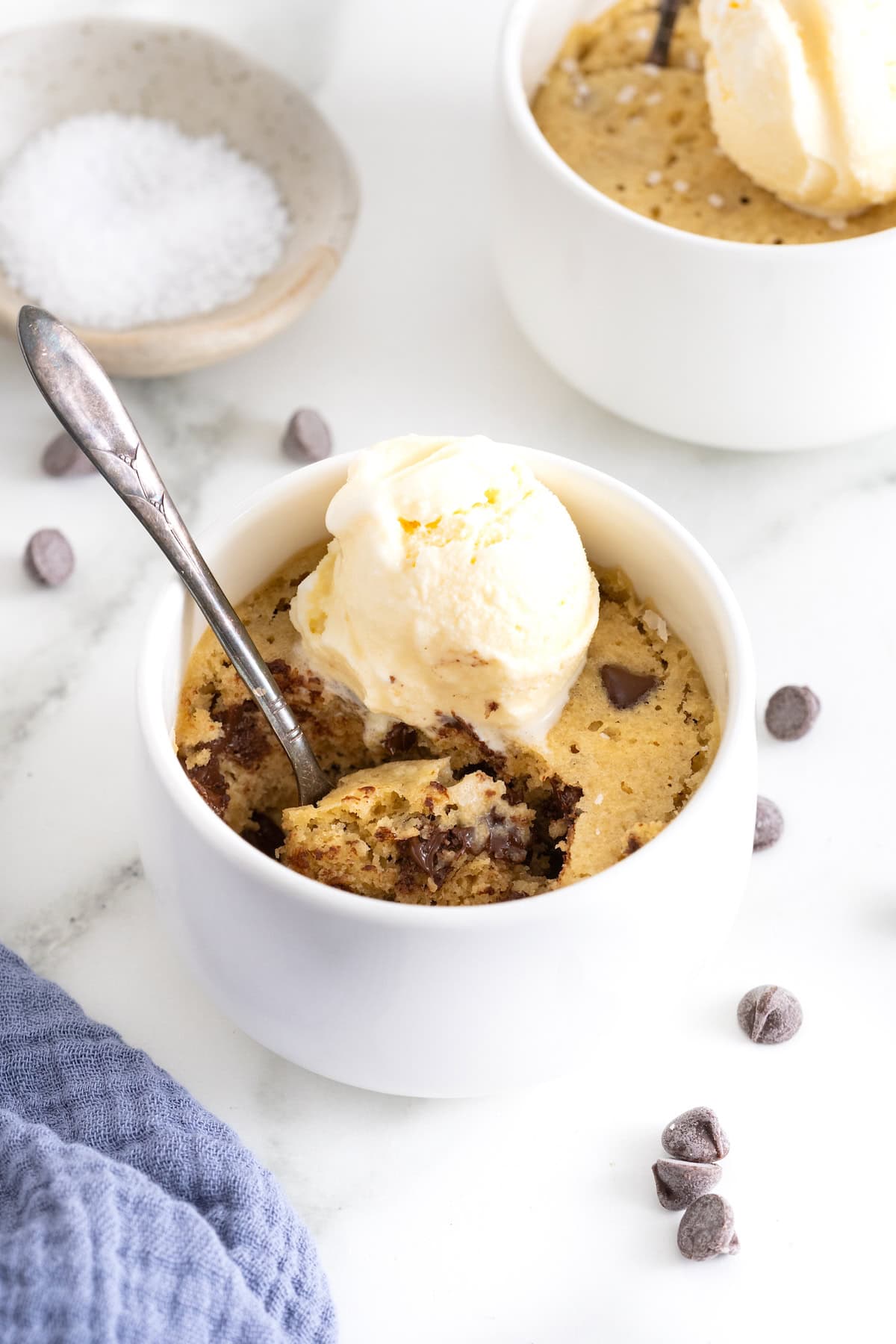 A microwave chocolate chip cookie in a white mug topped with a scoop of vanilla ice cream.