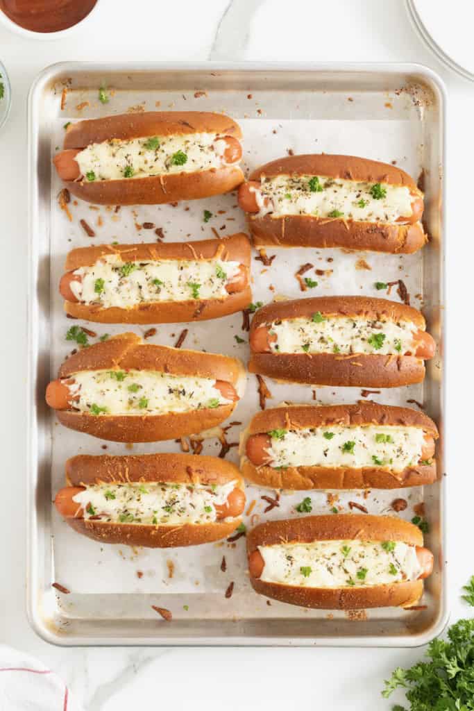 8 baked hot dogs with melted cheese and sprinkled with parsley on a parchment lined aluminum baking sheet.